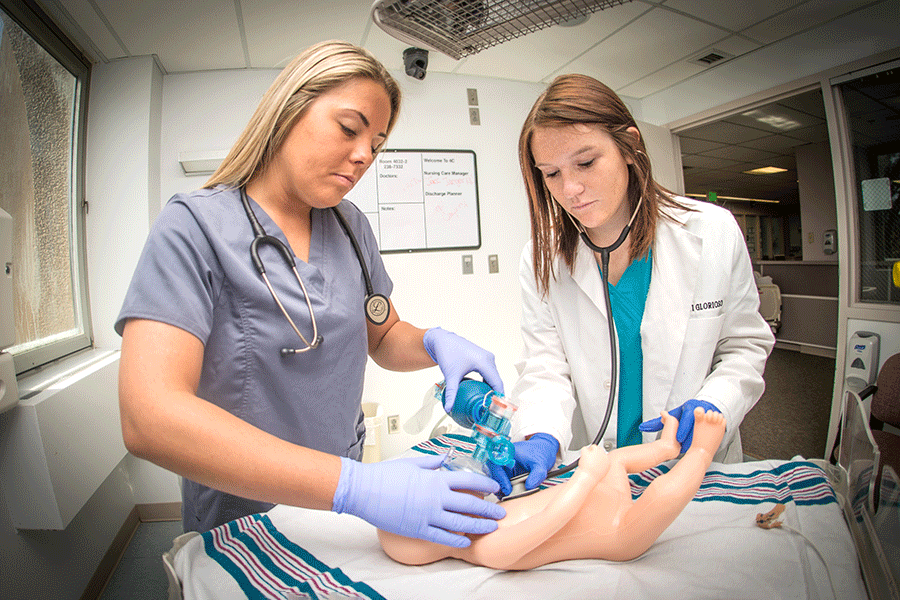 Two female students in a medical room. They are looking at an infant mannequin. On the right, a female student with brown hair wears teal scrubs, a white lab coat, and a stethoscope. On the left, a female student with blonde hair wears blue scrubs. She holds an oxygen mask to the mannequin.