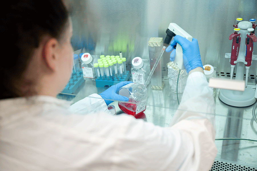 The lab tech faces away from the camera while using lab equipment to sample a substance in a container.