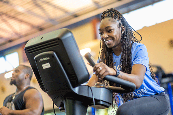 A smiling Black woman with long dreadlocks rides an exercise bike in the Student Recreation Center. She is wearing a light blue t-shirt and dark workout pants. Visible to her right is a smiling, muscular Black man with facial hair and wearing a black tank top. Daylight streams through elevated windows behind them, and yellow walls and a corrugated ceiling are also visible.