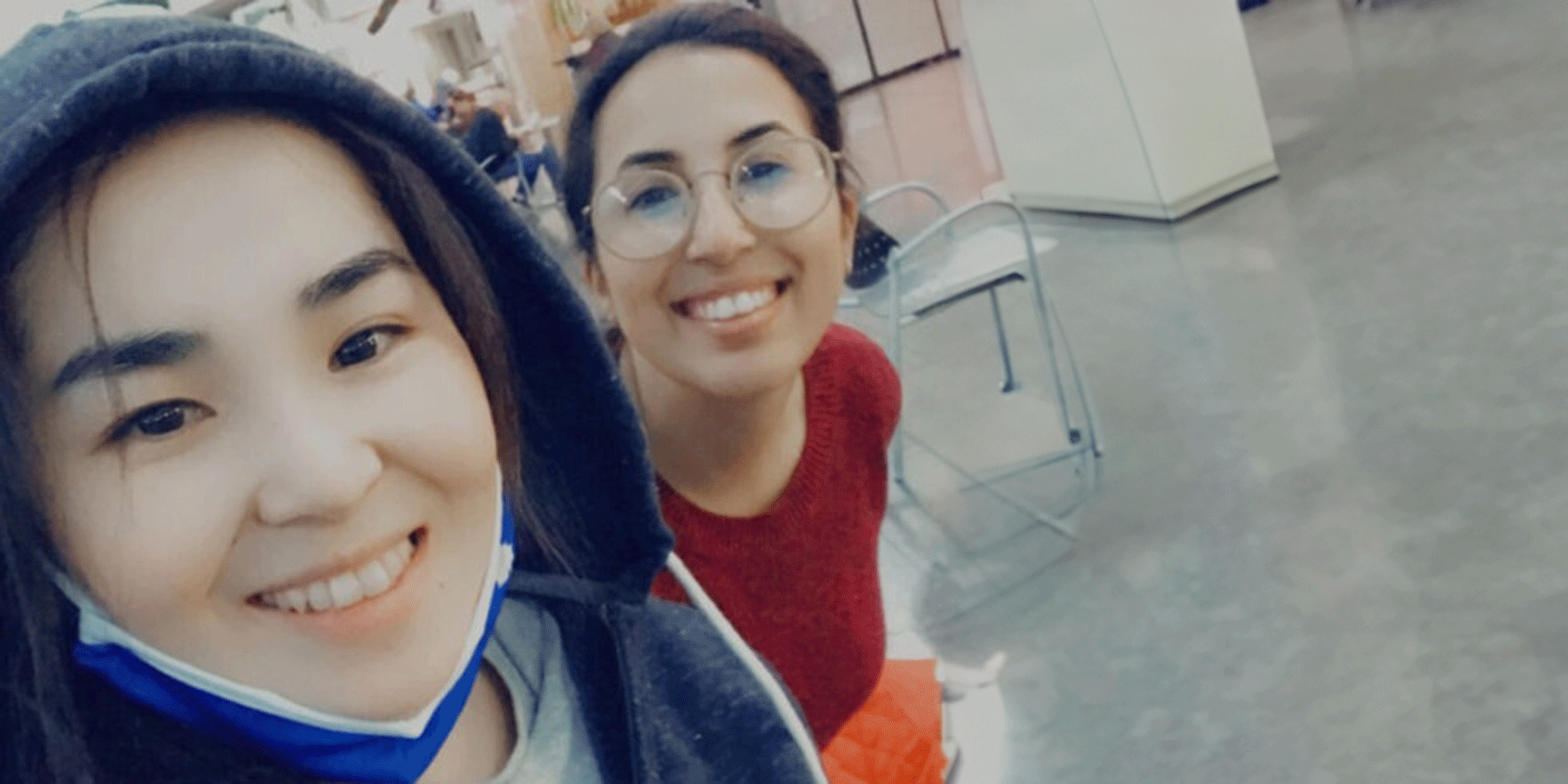 Two female students of Asian heritage pose for a selfie. On the left is a female student with brown hair. She wears a dark grey jacket with the hood up, and a blue mask pulled down from her mouth. On the right is a female student with brown hair pulled back. She wears glasses and a red sweater. Behind them are cabinets, a chair, and other individuals blurred in the background.