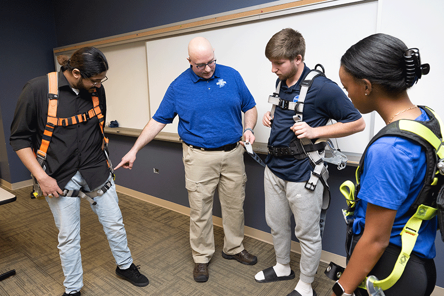 Four individuals are inside a classroom with a whiteboard behind them. On the left is a male student with darker skin. He has long dark brown hair pulled back into a ponytail. He wears a black shirt, denim pants, and an orange safety harness around his shoulders. To his right is a middle-aged white bald male, wearing black glasses, a blue polo shirt, and khaki pants. He is pointing to the student to his left. To his right is a white male student with short brown hair.