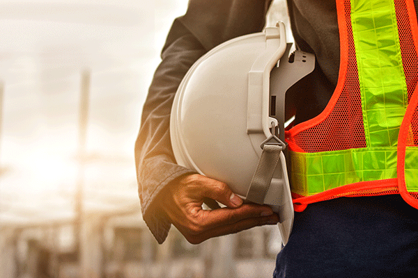 A person holding a white construction hat while wearing an orange-and-yellow safety vest over a grey long-sleeved shirt, and dark pants. A grey-and-white background with indistinct structures is blurred behind the individual.