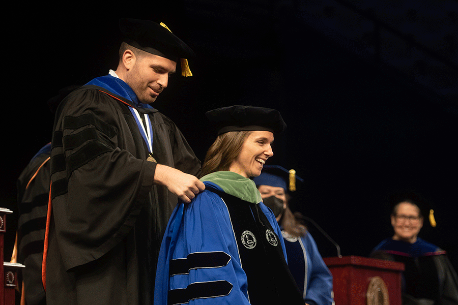 An individual in Commencement regalia fixes the hood of another individual in Commencement regalia