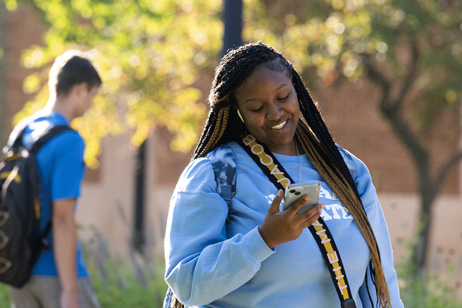A Black female student is standing outside, looking down at a smartphone in her right hand. She has long black and brown braided dreadlocks, and she is wearing a blue sweatshirt with Indiana State in white lettering on the front. A blue backpack is visible around her shoulders, and she is carrying a black-and-gold crossbody purse with white lettering on the strap. A white male student wearing a blue T-shirt is visible in the background. Trees and a brick building are also visible behind the female student.