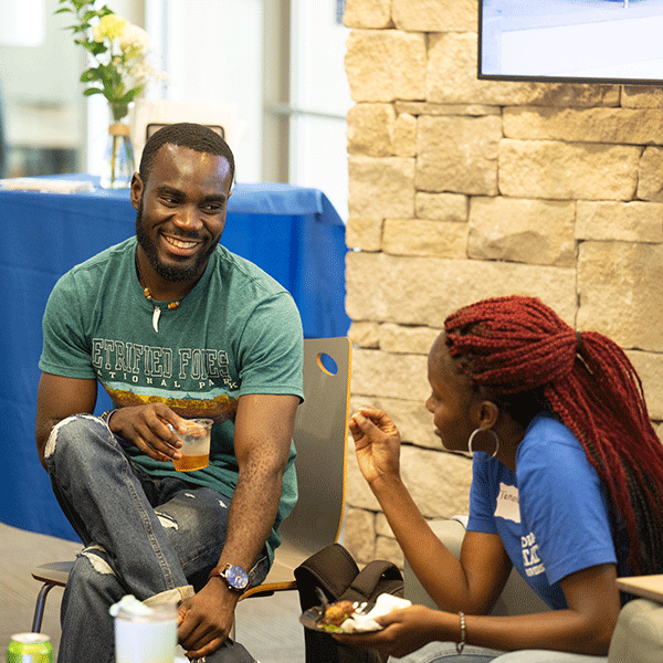  a Black male student wearing a green shirt smiling while talking to a black female student with red heard and a blue shirt in the welcome center. 