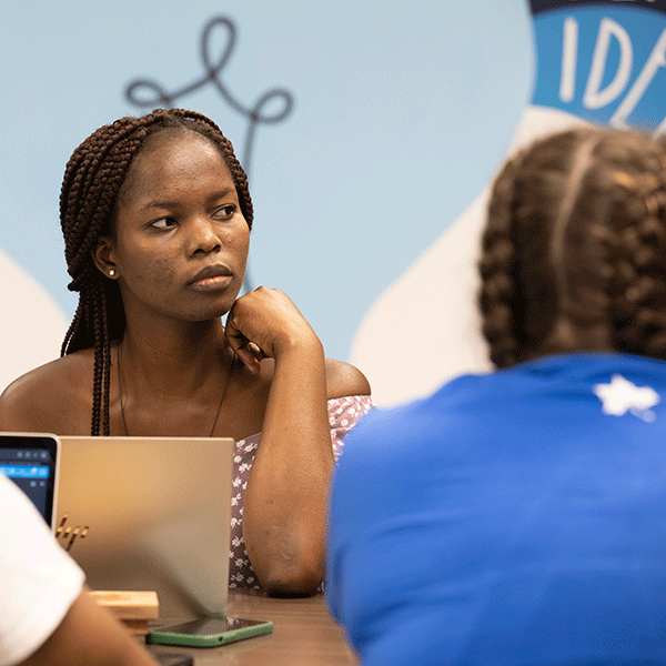 A Black female student listens intently and looks to the right while seated at a table with another student in the foreground with a blue shirt with back to the camera. 