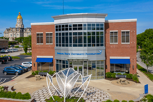 The image shows the Indiana State University Foundation building, which is modern and made of brick and glass. In front of the building, there’s a distinctive white sculpture that resembles an open sycamore leaf. The foundation building has large windows and is adorned with blue awnings over the entrances. There are several cars parked beside the building indicating it might be a busy day or time.