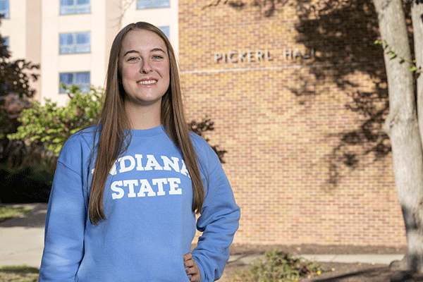 Portrait of a white female with long brown hair and wearing a light blue Indiana State long sleeved shirt looks at the camera with a brick building with “Pickerl Hall” on it in the background.