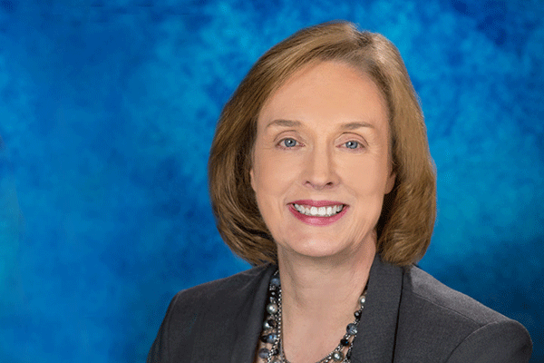 Portrait of a white woman with shoulder length brown hair smiling and looking into the camera with a blue background.