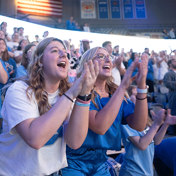 Two female sycamore fans cheer and clap and are surrounded by other cheering students in the fan section of a basketball game. 