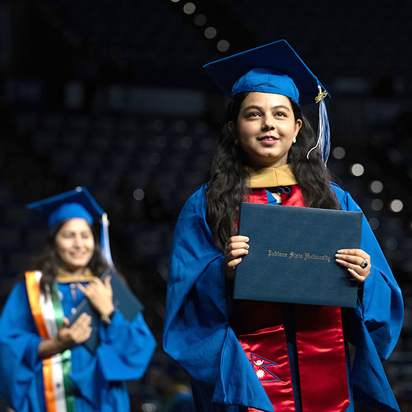 A female student with a blue graduation gown and wearing a graduation cap crosses the stage during commencement. 