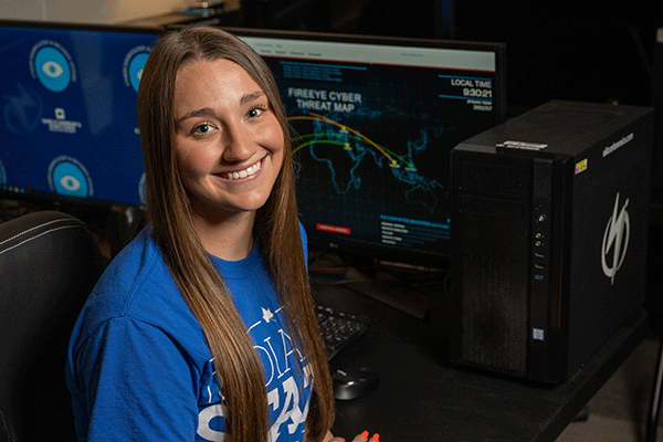 A white female student with long brown hear wearing an Indiana State shirt looks back at the camera while sitting in a dark room with a computer with a threat analysis website on the screen. 