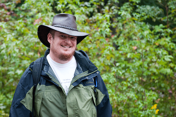 Portrait of a white male professor with mustache and beard wearing a hat and light jacket with green foliage in the background.