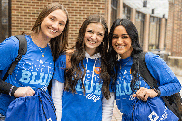 Three female students wearing blue Give to Blue shirts pose and smile