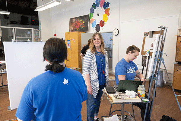 Two female students are seated in an art studio, and their female instructor stands between them. The instructor has dark blonde hair and is wearing a blue t-shirt with a grey-and-white striped, unbuttoned shirt over top of it, blue jeans, and a blue-and-white lanyard with something red hanging from the end. She is smiling at the student in the foreground, who has short back hair and whose back is turned to the camera as she sits in front of her canvas. The other student, pictured to the instructor’s left.