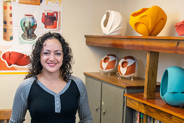 A smiling Latina woman with curly black hair and wearing a black-and-grey, long-sleeved jersey t-shirt with buttons stands in a room with colorful pottery pieces on shelves beside her. Visible behind her is a poster that illustrates examples of other pottery designs.