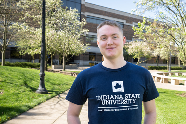 A white, male student smiles as he stands at some distance in front of a multi-story brick building, the John T. Myers Technology Building. The student has short blond hair and is wearing a navy blue t-shirt that says “Bailey College of Engineering & Technology” on it. It also has the blue shape of a Sycamore leaf logo in a white box. In the background, green grass and trees with white flowers can be seen.