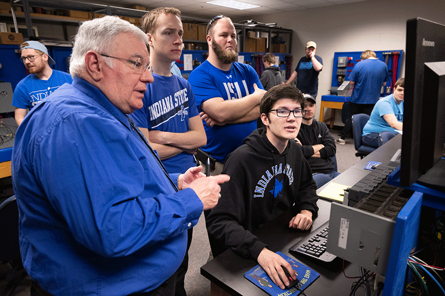 A white, older professor with white hair and glasses, and wearing a blue windbreaker jacket, stands next to two white male students in a control room as a third white male student sits at a computer terminal. The two standing students have short, blondish-brown hair and glasses and are wearing blue Indiana State t-shirts. The one further away from the instructor also wears a beard. The seated student has dark hair and glasses and wears a black Indiana State hoodie with blue letters on it.