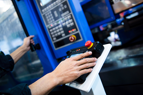 A pair of hands are shown engaging with a blue control panel. The right hand rests on a black control pad with two illuminated orange lights and a red button on a yellow base, and a few other buttons and lights on the far side. The pad sits on a white pedestal. In the background, a tall blue control panel with various buttons, lights, and a red knob is also visible. The left hand in the image rests further away on a black vertical handle attached to the control panel.
