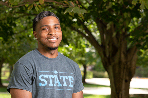 A smiling Black male student with black hair stands outside on campus with green, shady trees and sunshine in the background. He wears a grey shirt with STATE on it in light blue.