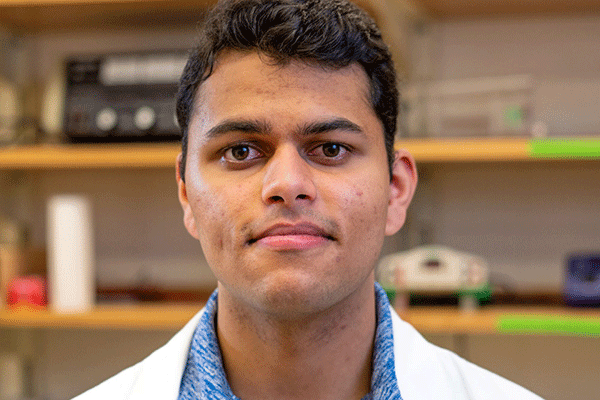 A close-up picture of a male student of Indiana heritage staring directly into the camera with a soft smile. He is wearing a blue shirt and white lab coat. 