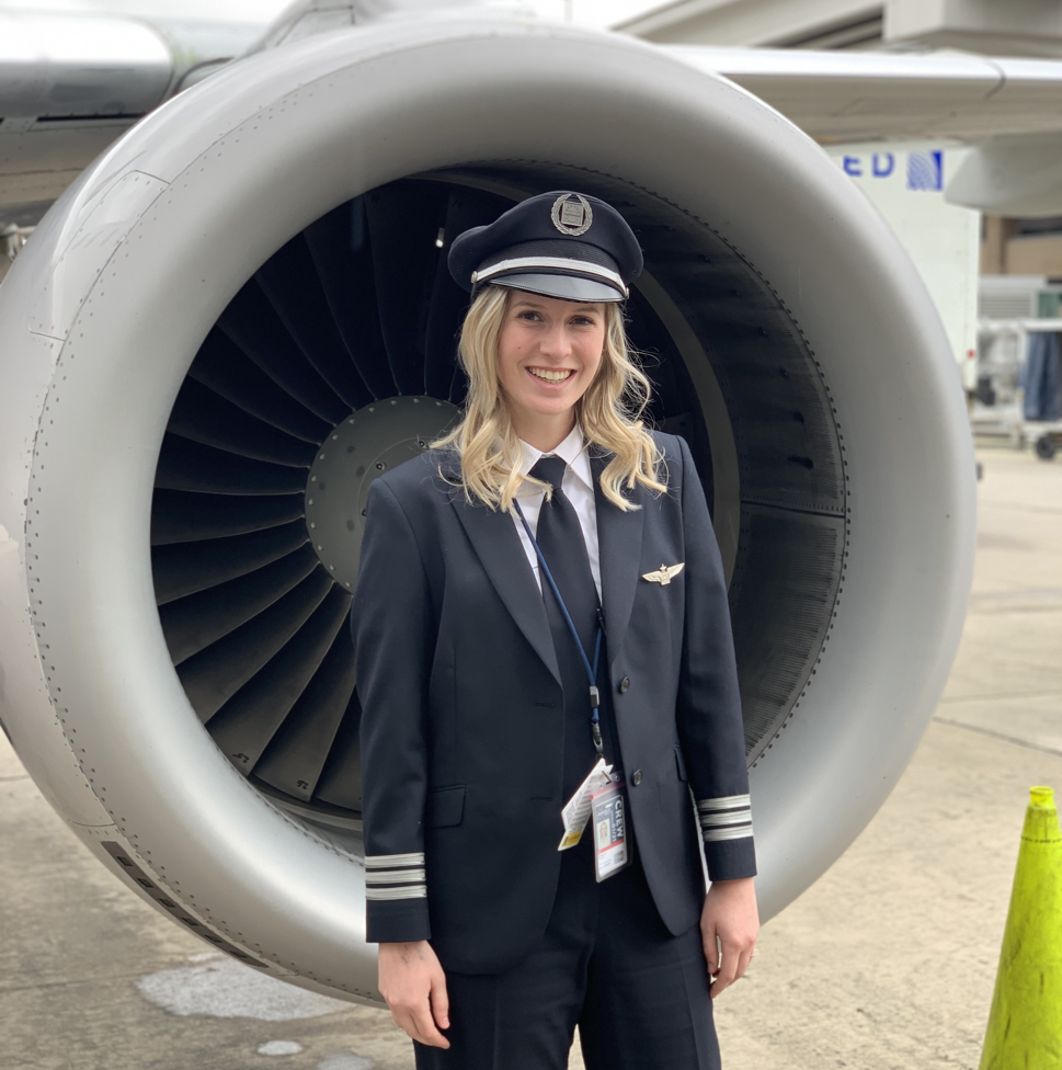 A smiling young blonde woman in a black and white pilot’s uniform and cap stands in front of an airplane’s silver-and-gray turbine engine.  