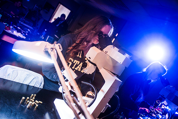 female student looking into a microscope in a darkly lit criminology lab with blue light in the background, and spent bullets on the table on the left.