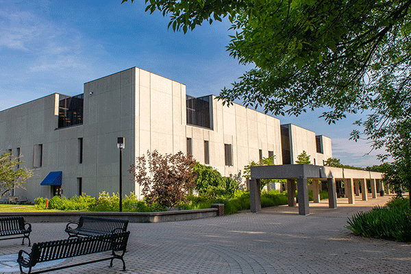 white, multi-story square building in the distance with meal benches surrounding an ISU seal on the ground and a walkway leading to the building. A tree with green leaves is on the right with blue sky above.