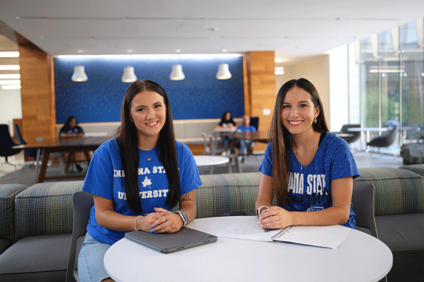 Two white female students wearing blue shirts smile while sitting at a table in the Health and Human Services building.