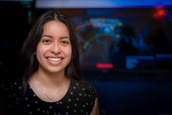 A young Latina woman with long dark hair smiles at the camera in a computer lab. The woman is wearing a black shirt with a white pattern and a silver necklace. A video monitor showing a colorful map of simulated cyberattacks is blurred but visible behind her.