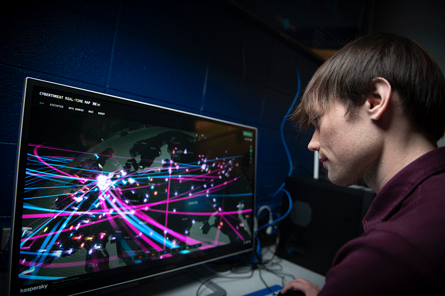 A white male student with short brown hair and wearing a black shirt is working on a computer. The monitor displays a colorful map of simulated cyberattacks. 