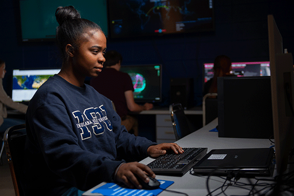 Female student of color with black hair in a bun and wearing a navy Indiana State University sweatshirt works on computer in a dark computer lab. Illuminated computer terminals and large-screen monitors are visible behind her.