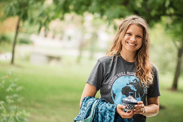 A white female student with long curly light brown hair poses outside. She wears a grey T-shirt with a globe showing North and South America in blue colors. “Where Will Blue” in white lettering is visible above the globe. She holds a blue jacket in her right hand and holds a blue-and-white ceramic cup with a lid in her left hand. Green trees are visible in the background, blurred in the photo. 