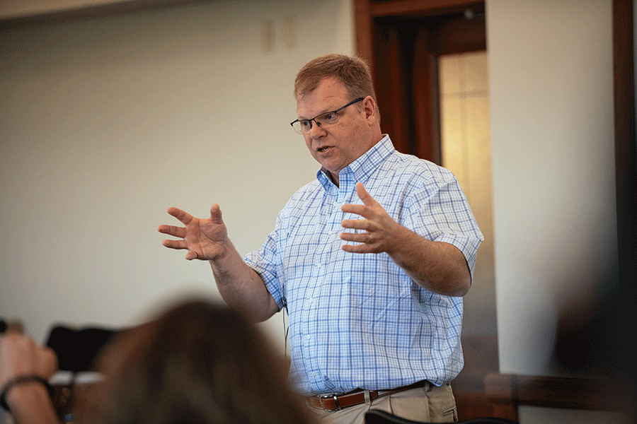 A white middle-aged male with short brown hair stands in a room, talking to a group of individuals blurred in the photo. He wears glasses, a white-and-blue checkered dress shirt, and khaki dress pants. Both of his hands are raised as he talks. 