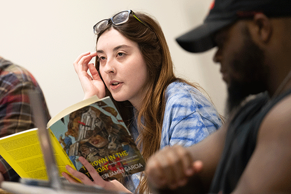 A white female student holds a book and looks to the left in a classroom with a Black male sitting to the right of her.