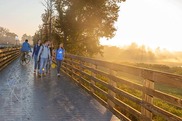 A group of four students walking along a wooden boardwalk that overlooks a scenic open area of marshland in early morning as the sun breaks through the clouds.