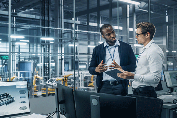 Two men stand in a manufacturing testing facility. They are looking at each other and appear to be trying to solve a problem. On the left is a Black male with black hair and a short beard, wearing a light blue, open-collared shirt, a navy blue jacket, dark blue pants with a black belt, and a blue lanyard around his neck. He holds a pencil in his left hand, and both hands are raised slightly and open in toward himself On the right stands a white man with dark brown hair and glasses.