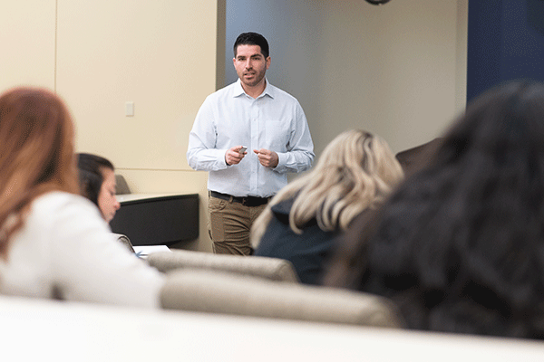 An adult white male teacher with short hair, standing in the background, focused and teaching a class. In the foreground, the blurry backs of students' heads are visible, emphasizing the teacher's engagement with the class.