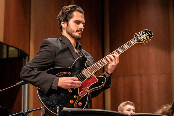 A male with a black shirt and dark hear with beard and a mustache plays a black guitar.  