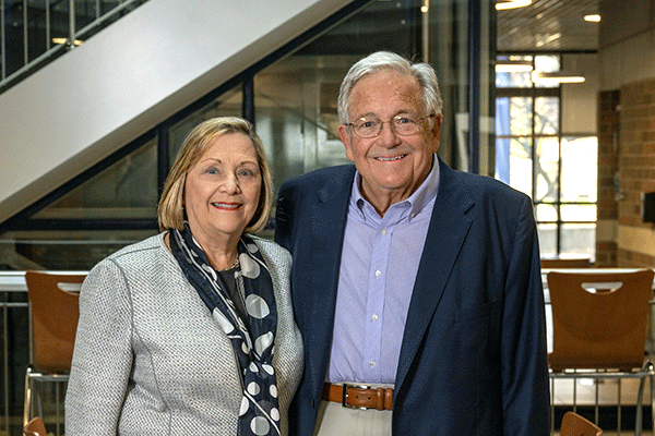 Gloria (left) and Steve (right) Bailey stand smiling in the Bailey College of Engineering & Technology Building. She is wearing a beige business casual suit with a black scarf that has large white circles on it. He is wearing a dark blue sport coat with light blue shirt, beige khakis, and brown belt. The visible interior of the building is dark, with outdoor sunlight visible through windows in the back of the building