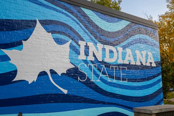 Large mural with a white Sycamore leaf and “Indiana State” written in white text with various shades of wavy blue lines as the background on the left side of the image while on the right is a black lamp post with the light on with trees with yellow fall leaves behind. 