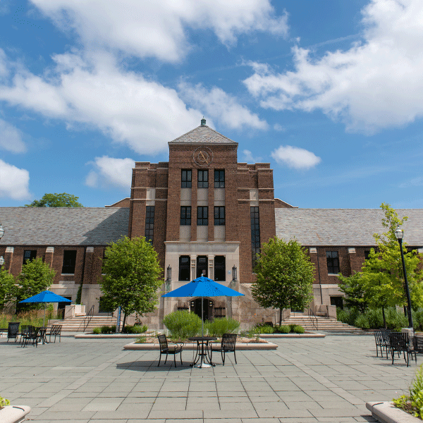 The exterior of a multi story brick building with a patio in front, and la large staircase on the left and right side leading up to the doorway. The sky is blue with white fluffy clouds, and there is a clock tower near the top of the building.
