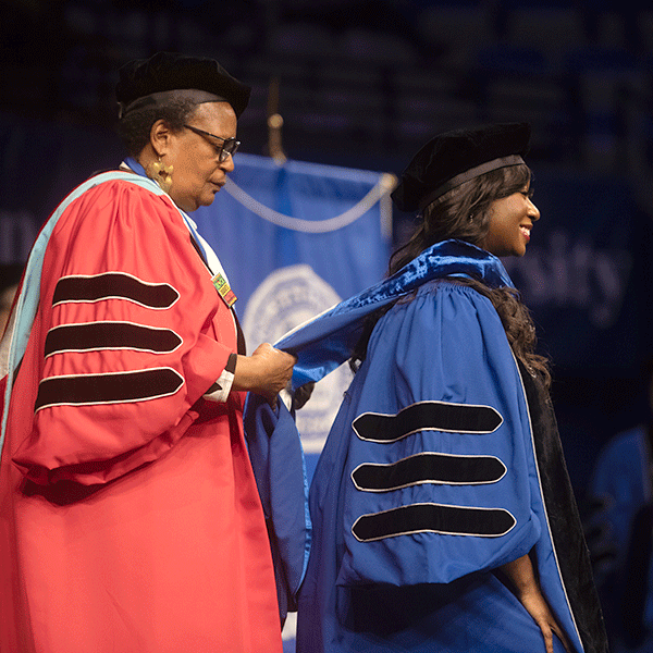 A Black female professor wearing a red commencement robe, places doctoral hood over the head of a Black female student during a commencement ceremony for Graduate Students.