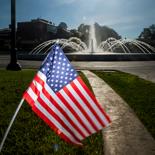An American flag with white stars on a blue background, and red and white stripes in front of a fountain with one spout in the center and several small spouts around the center in a circular manner.