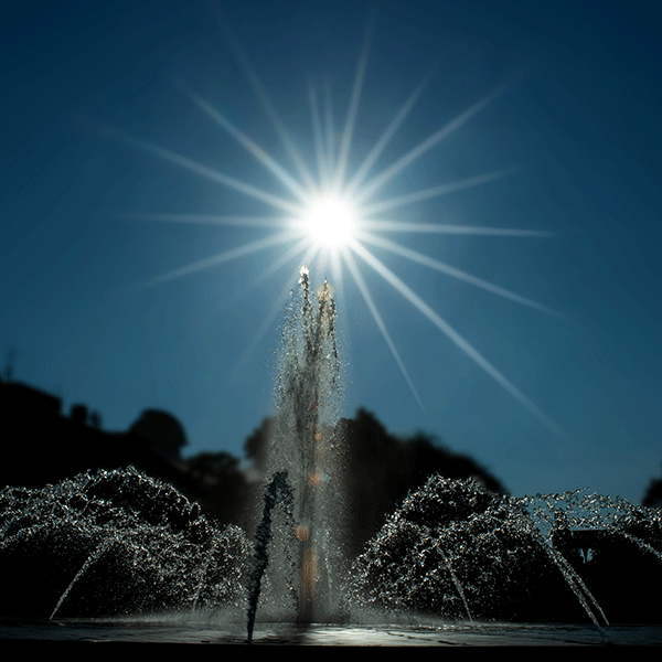 Darkly lit photo of a fountain with a center spout and smaller spouts around the center in a circular pattern with the sun behind with sunbeams extending in all directions from the center of the sun.