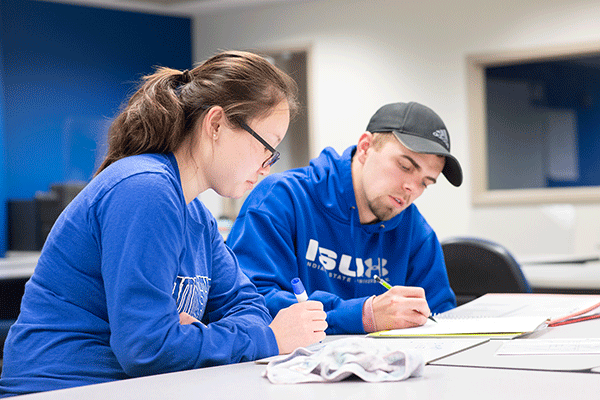 Two students, an woman with dark hair in a ponytail and glasses, and a man wearing a grey baseball cap, work on an assignment together. They both wear blue ISU apparel, and both hold writing implements in their hands.