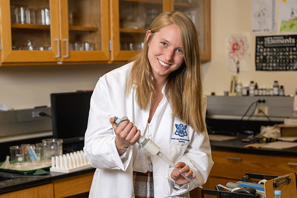 A white female student in a white lab coat looks towards the camera in a lab.