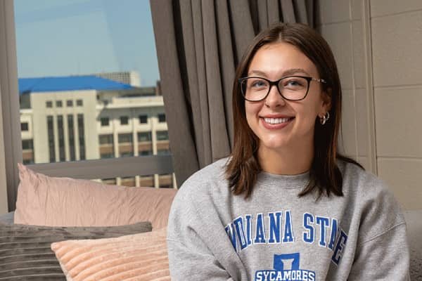 A white female student with shoulder-length brown hair is sitting on a bed inside a dorm room. Pink and grey pillows are visible on the bed. The student is wearing dark glasses and a grey sweatshirt with Indiana State University in blue lettering, along with the blue Sycamores athletics logo. Behind her, a window with open grey curtains shows a view of a concrete building.