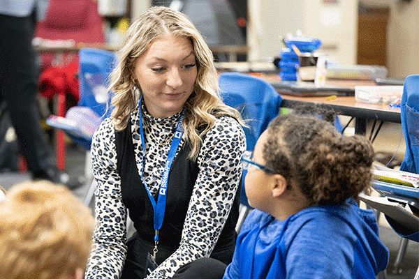 A white female student with shoulder-length curly blond hair sits in a classroom. She wears a cheetah-print, black-and-white, long-sleeved shirt, a black vest, and a blue lanyard. On her right is a young Black female child with brown hair pulled back into a bun. She wears a blue hoodie. Another child’s blond hair is visible to her left, and tables and blue chairs are visible in the background. 