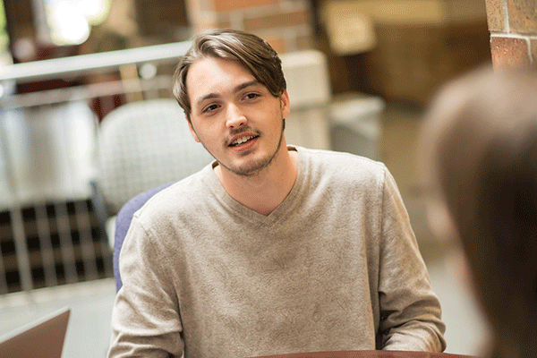 A smiling white male student with sparse facial hair and dark brown hair that falls left across his forehead is seated and talking to someone who is only visible from the back and is blurred. The man wears a beige sweatshirt. Behind him, out of focus, can be seen other tables, chairs, a brick pillar, a metal railing, and other elements of the commons area in the student union.